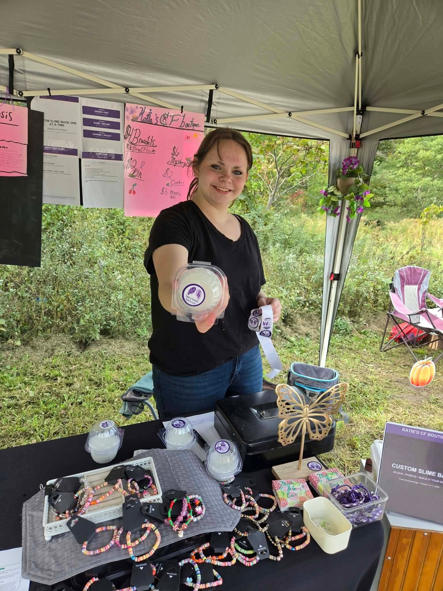Katie holds up a cup of custom slime from Katie's CF Boutique, surrounded by handmade bracelets and charms, at the Creek Rd Kids Bridge