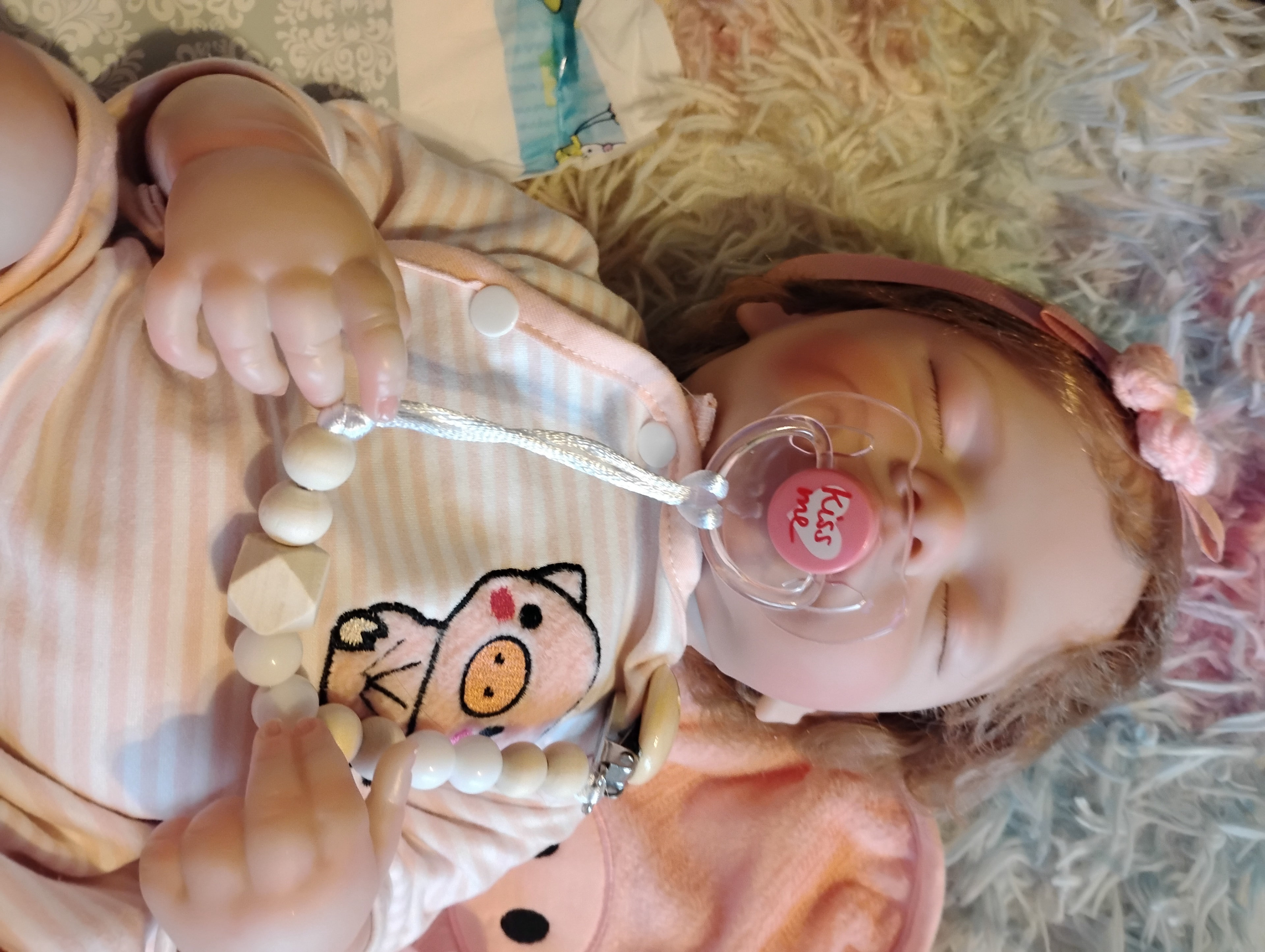 Baby doll donated by Bill and Clara Lundgren to the Kids Care Crew, holding a wooden-bead pacifier clip and dressed in a pink pig-themed outfit with a heart pacifier