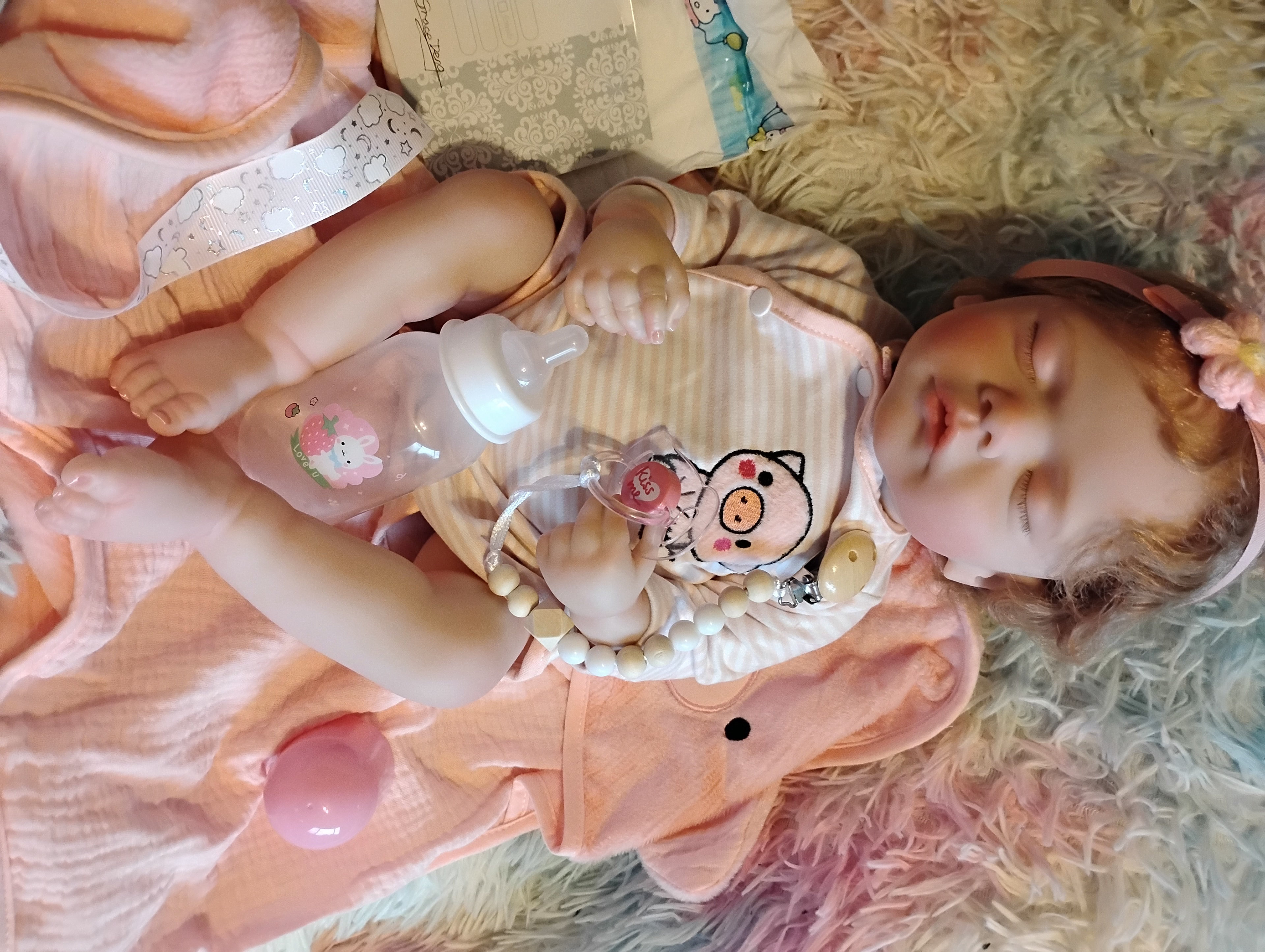 Baby doll donated by Bill and Clara Lundgren to the Kids Care Crew, resting on a pink pig-themed blanket with a bottle, pacifier, and keepsake ribbon